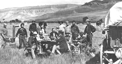 A group of men gathered around a table outdoors, with mountains in the background and a wagon nearby.