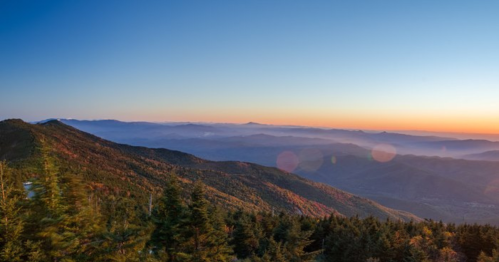 A panoramic view of mountains at sunset, with layers of hills and a clear sky transitioning from blue to orange.