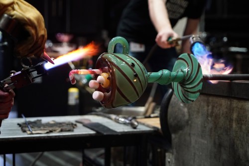 A glassblower uses a torch to shape a colorful glass piece with intricate details in a workshop setting.
