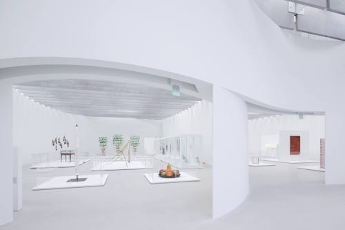A spacious, modern art gallery with white walls, showcasing various sculptures and installations in glass displays.