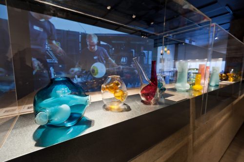 Colorful glass vases displayed in a gallery, with a video of glassblowing in the background.