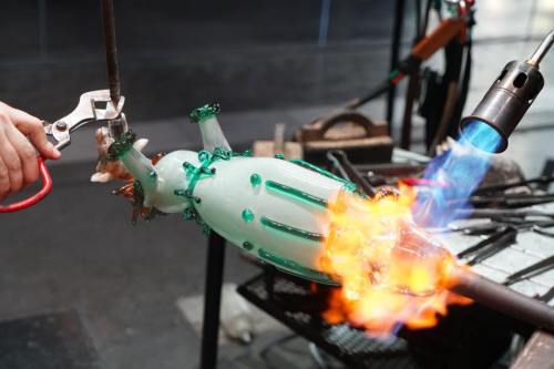 A glassblower shapes a green glass sculpture with a torch, creating flames and intricate details.