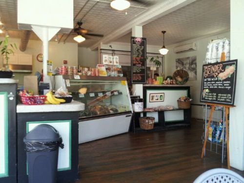 Cozy deli interior with a display case, menu board, and seating area, featuring plants and a trash can.