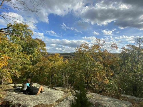 A person sits on a rock, overlooking a vibrant autumn landscape with colorful trees and a cloudy sky.