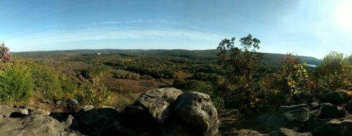 Panoramic view of a colorful autumn landscape with rolling hills, trees, and a clear blue sky. Rocks in the foreground.