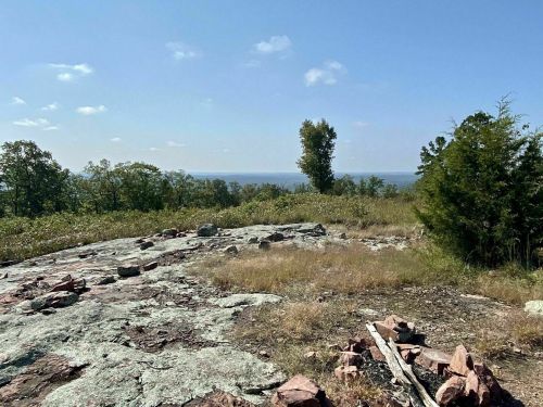 A rocky landscape with sparse grass and trees, overlooking a distant body of water under a clear blue sky.