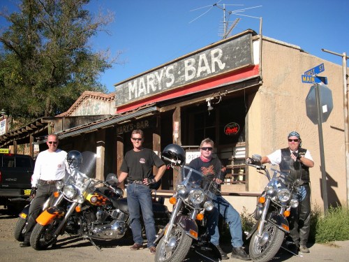 Four motorcyclists pose in front of "Mary's Bar," with vintage motorcycles parked nearby under a clear blue sky.