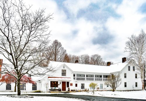 A white, two-story house surrounded by snow, with bare trees and a cloudy sky in the background.