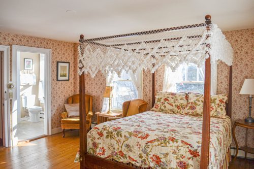 Cozy bedroom with a four-poster bed, floral bedding, and vintage decor, featuring a bathroom visible in the background.