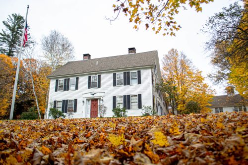 A white colonial-style house surrounded by colorful autumn leaves and trees, with an American flag in the foreground.