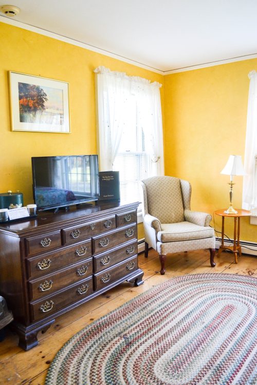 Cozy room with yellow walls, a patterned rug, a wooden dresser, a TV, an armchair, and a lamp by the window.