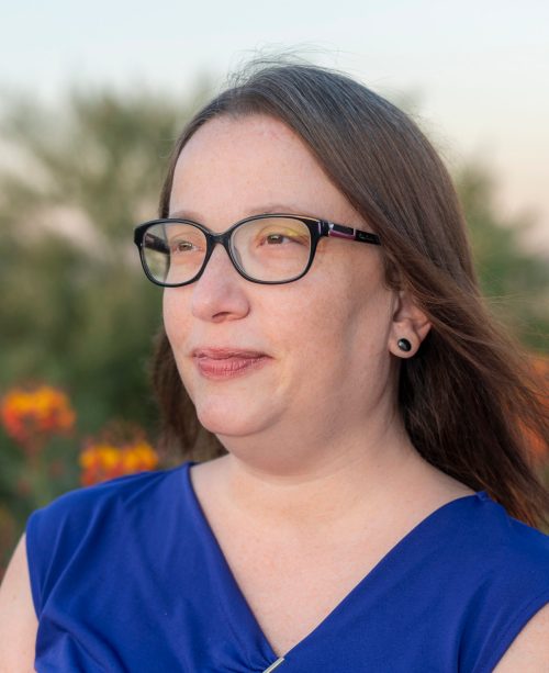 A woman with glasses and long hair smiles softly, wearing a blue top, with flowers and greenery in the background.