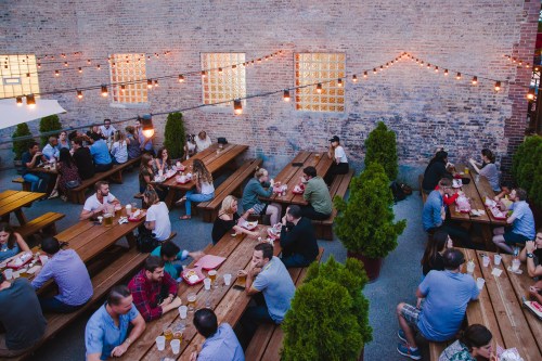 A lively outdoor dining area with people seated at wooden tables, string lights overhead, and greenery around.