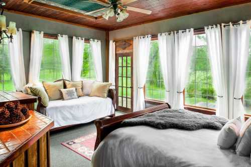 Bright, airy bedroom with large windows, a cozy bed, and a seating area, surrounded by greenery.