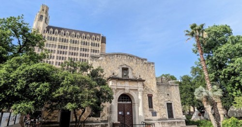 Historic building with ornate entrance, surrounded by trees, with a tall modern building in the background under a clear sky.