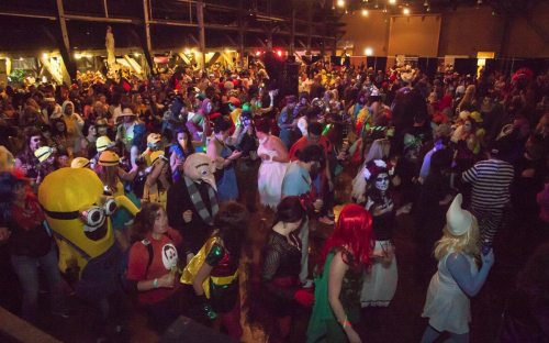 A vibrant crowd in costumes at a lively event, with colorful lights and various characters celebrating together.