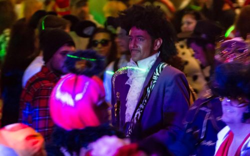 A crowd of people in colorful costumes, with one man in a purple outfit and curly hair standing out.