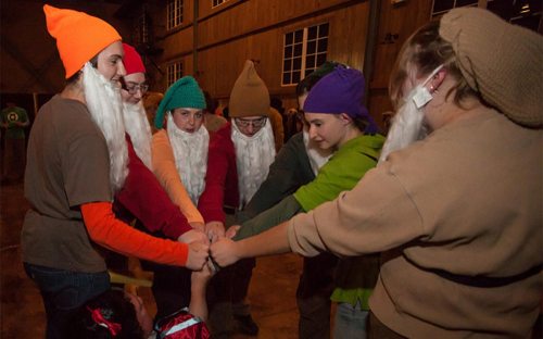 A group of people dressed as gnomes with colorful hats and white beards, huddled together in a playful pose.