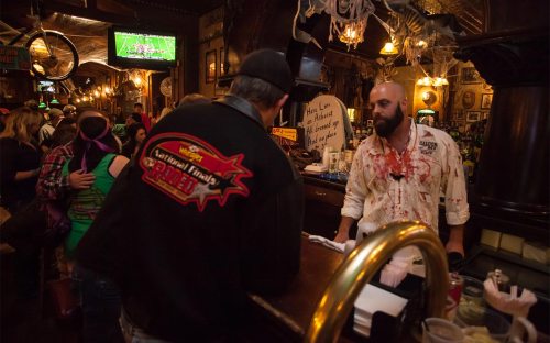 A bar scene with patrons, a bartender in a blood-stained shirt, and Halloween decorations in a lively atmosphere.