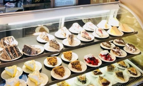 A display case filled with various cakes and desserts on white plates, including slices of pie and whipped cream toppings.