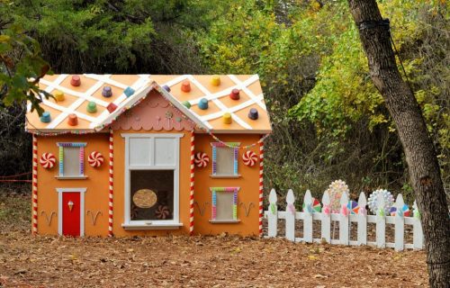 A whimsical gingerbread house with colorful decorations and a white picket fence, surrounded by trees.