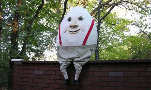 A large, cartoonish egg character with a smiling face, wearing red suspenders and beige pants, sitting on a brick wall.