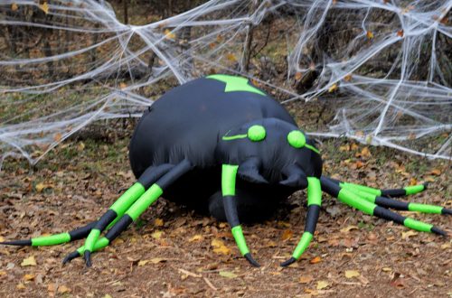 A large inflatable black spider with green accents sits on the ground, surrounded by fake cobwebs and fallen leaves.