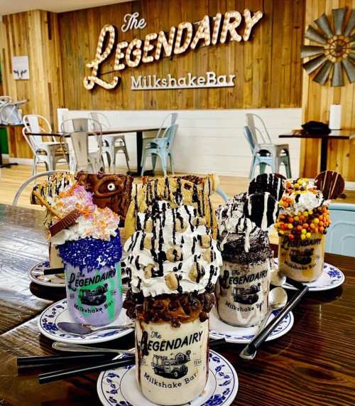 Five extravagant milkshakes topped with whipped cream, candies, and cookies, displayed on a wooden table.
