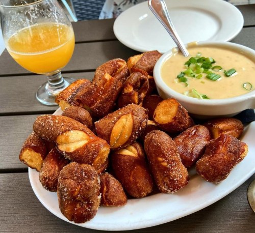 A plate of golden-brown pretzel bites with a side of creamy dip and a glass of orange drink on a wooden table.