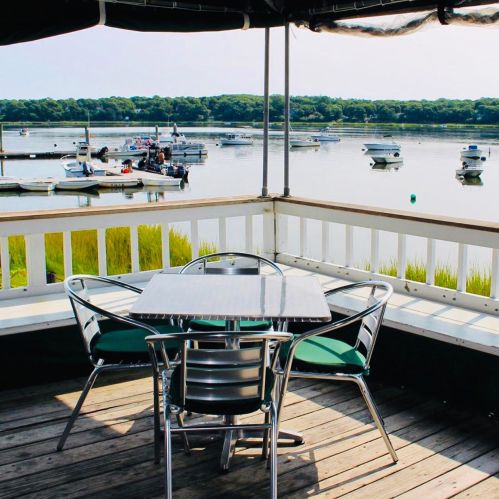 A waterfront view from a deck with a table and chairs, overlooking boats on calm water and lush greenery in the background.