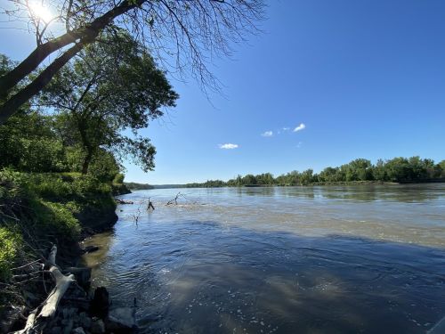 A serene river scene with trees along the bank under a clear blue sky and a few clouds.