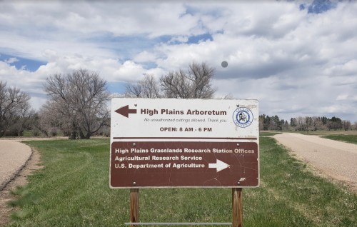 Sign for High Plains Arboretum with directions and hours of operation, set against a cloudy sky and gravel road.