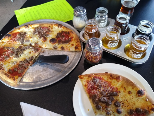 A pizza with sausage slices on a metal tray, alongside a plate with a slice, and a flight of craft beers in jars.