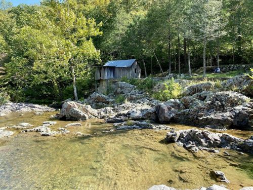 A rustic wooden cabin by a clear stream, surrounded by lush greenery and rocky terrain.