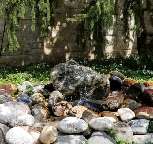 A serene rock garden with a large stone surrounded by smooth pebbles and gentle water flow, framed by greenery.