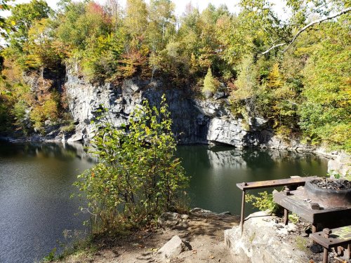 A serene lake surrounded by colorful autumn foliage and rocky cliffs, with a small grill in the foreground.