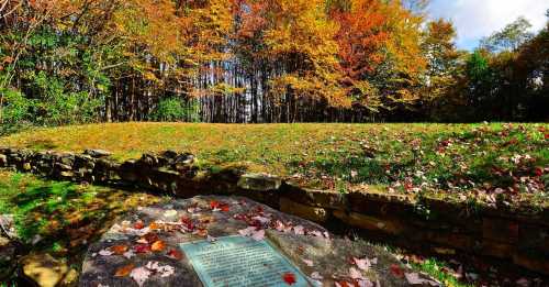 A stone plaque surrounded by colorful autumn foliage and trees, with fallen leaves on the ground.