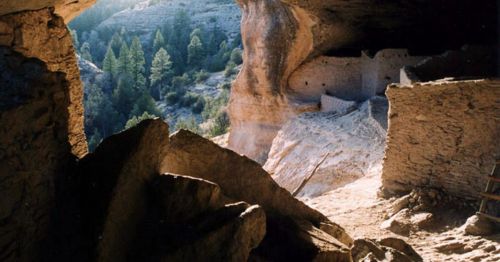 A cave entrance reveals ancient stone structures nestled in a forested landscape, illuminated by soft sunlight.