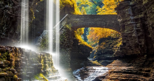 A serene waterfall cascades over rocks beneath a stone bridge, surrounded by vibrant autumn foliage.
