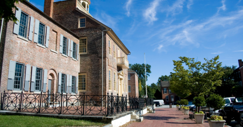 Historic brick building with large windows, surrounded by trees and a paved walkway under a clear blue sky.