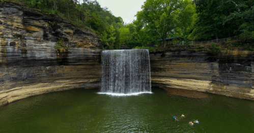 A serene waterfall cascades into a green pool, surrounded by rocky cliffs and lush trees. Two people swim nearby.