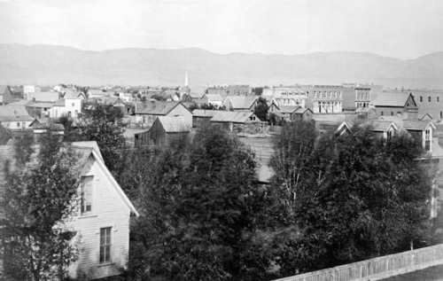 Black and white photo of a town with wooden houses and trees, set against a backdrop of distant mountains.