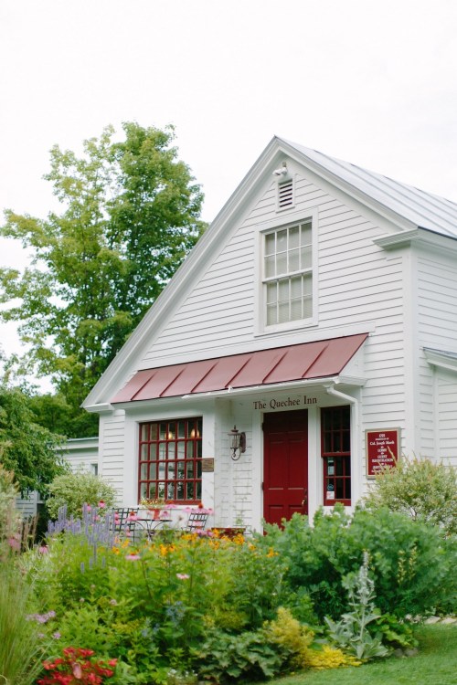 A charming white inn with a red door and roof, surrounded by colorful flowers and greenery.
