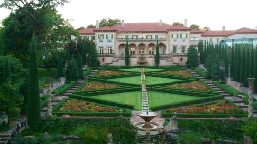 A beautifully landscaped garden with geometric flower beds and a fountain, set in front of a grand mansion.