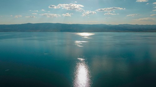 A serene landscape featuring calm water reflecting sunlight, with distant mountains and a partly cloudy sky.