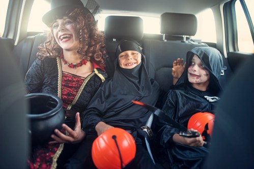 A woman in a costume and two kids in ninja outfits sit in a car, smiling and holding Halloween candy buckets.