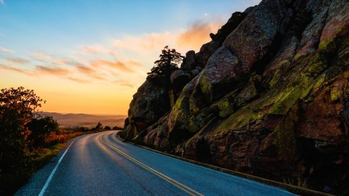 A winding road curves alongside a rocky landscape at sunset, with vibrant clouds in the sky.