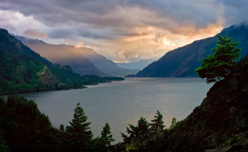 A serene river flows between mountains under a cloudy sky, with trees framing the foreground.
