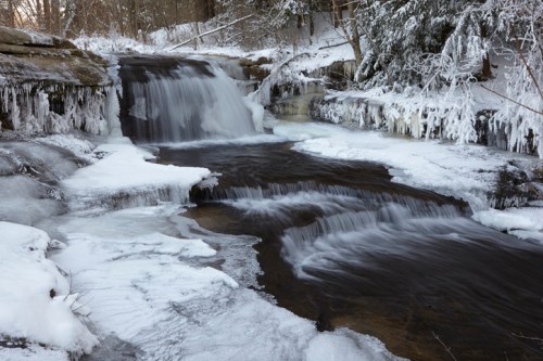 A serene winter scene of a flowing waterfall surrounded by ice and snow-covered rocks in a tranquil forest setting.