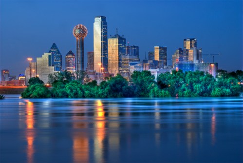 Dallas skyline at dusk, featuring illuminated buildings and reflections on the water. Lush greenery in the foreground.